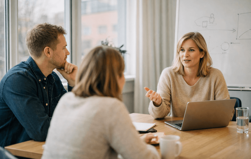 Tre kollegor i ett ljust mötesrum diskuterar kompetensutveckling för löneavdelningen, med laptop och anteckningsblock på bordet.